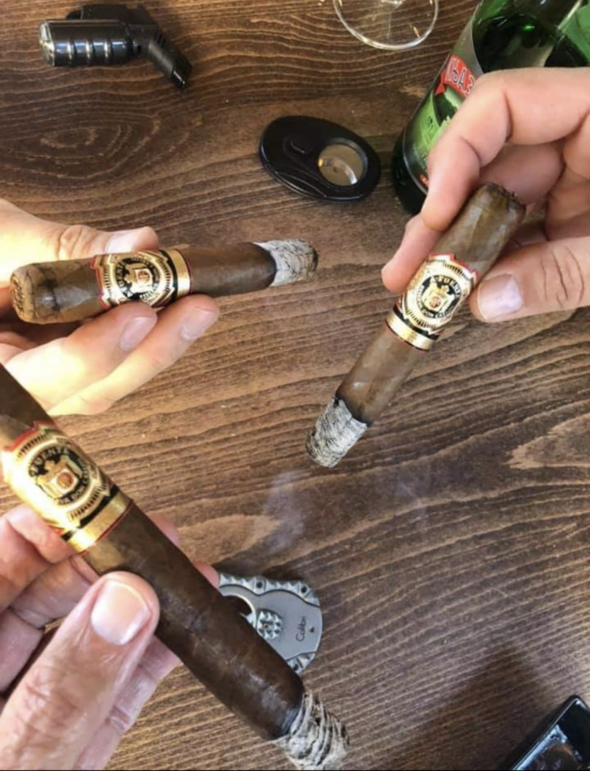 Three Arturo Fuente cigars resting on a dark wood table with a guillotine cutter and lighter.