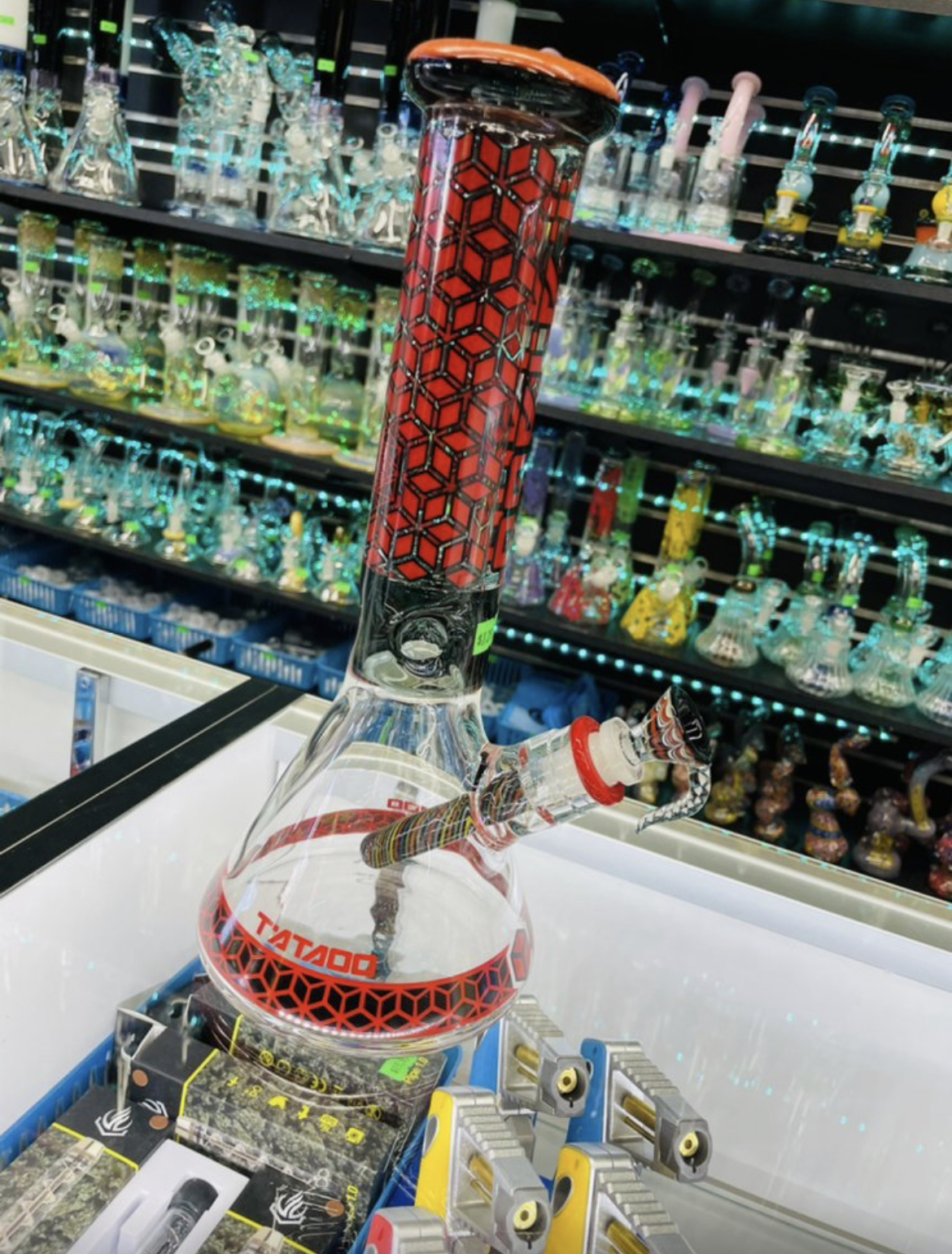 A red and black geometric-pattern Tataoo beaker bong glowing in front of a wall of glass pipes.