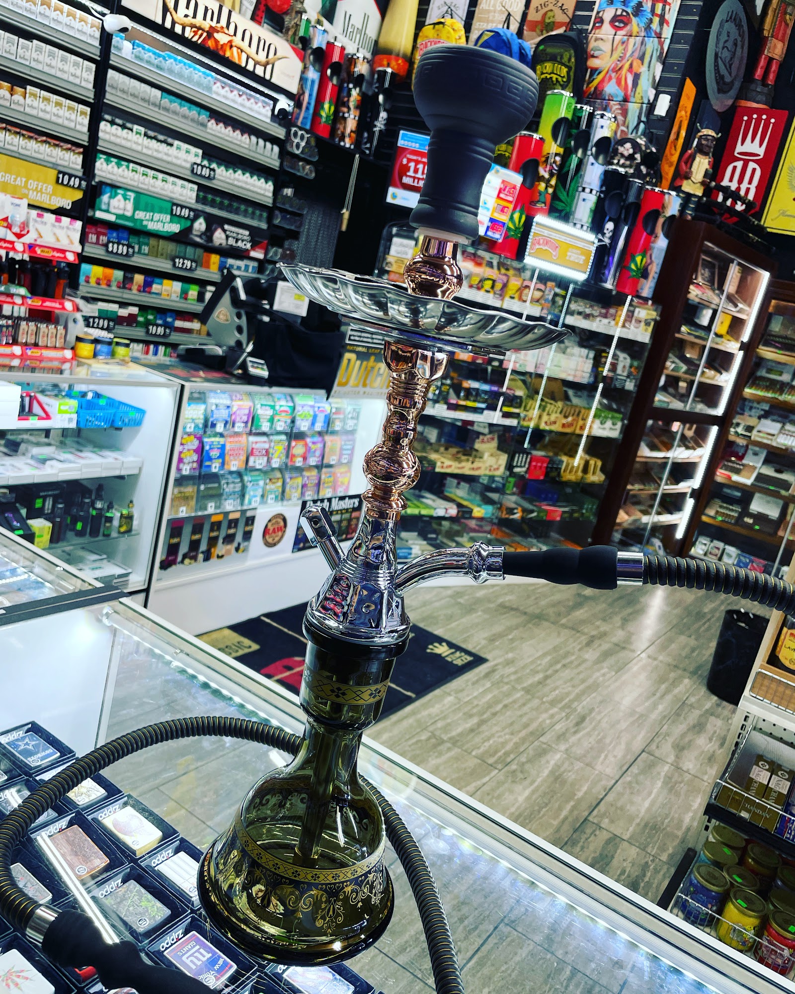 A tall ornate hookah with brass stem on a glass counter, with vape and accessory shelves visible behind.