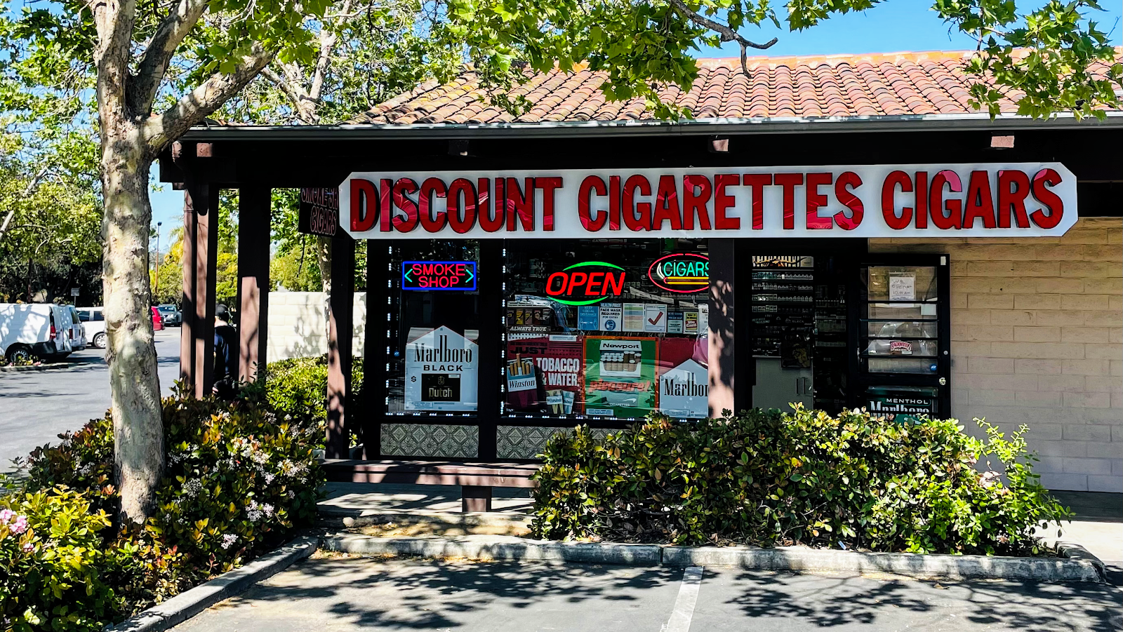 The Spanish tile-roof storefront of Discount Cigarettes Cigar on El Camino Real, with neon OPEN, CIGARS, and SMOKE SHOP signs in the window.